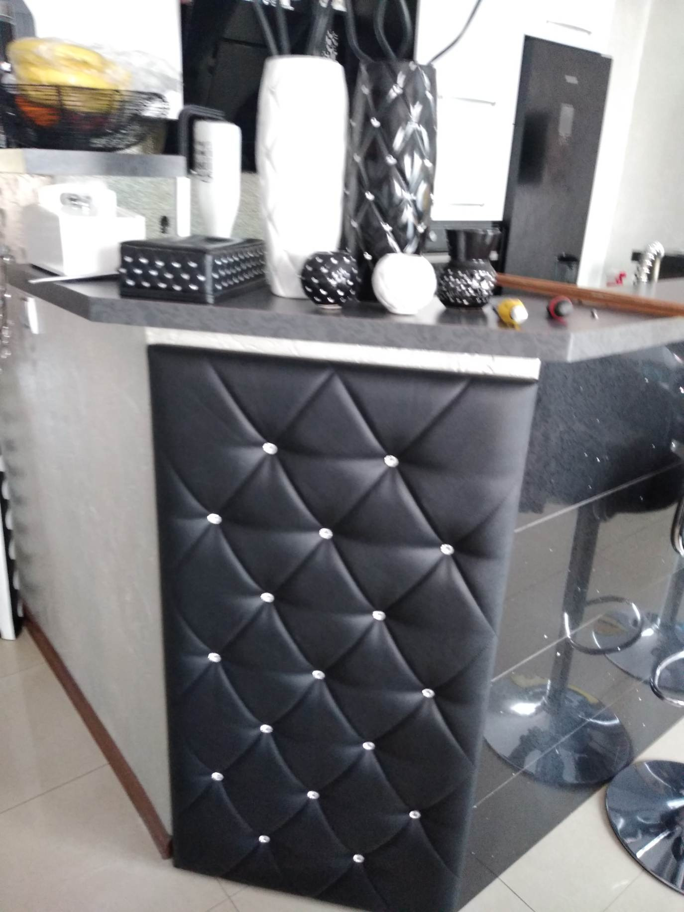 Wykończenie baru z czarnej pikowanej skóry z dekoracyjnymi kryształkami, widoczne również czarne hokery i dekoracje na blacie.