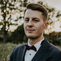 Portret młodego mężczyzny w garniturze i bordowej muszce na tle rozmytej zieleni, delikatny uśmiech.