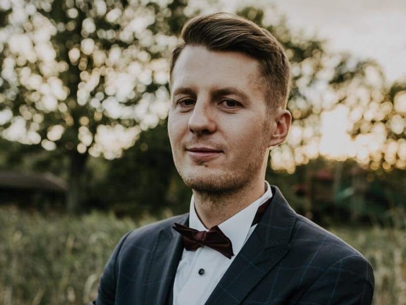 Portret młodego mężczyzny w garniturze i bordowej muszce na tle rozmytej zieleni, delikatny uśmiech.