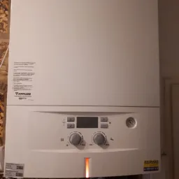 Biały piec gazowy Junkers z wyświetlaczem i regulacją, zamontowany na ścianie z widocznymi rurami miedzianymi w dolnej części.