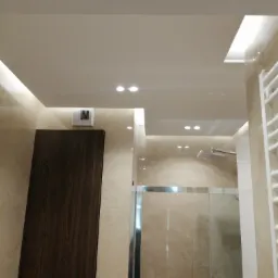 Wykończona łazienka z beżowymi płytkami, kabiną prysznicową ze srebrnymi profilami, drewnianą szafką i podświetlanym sufitem podwieszanym z oświetleniem punktowym.