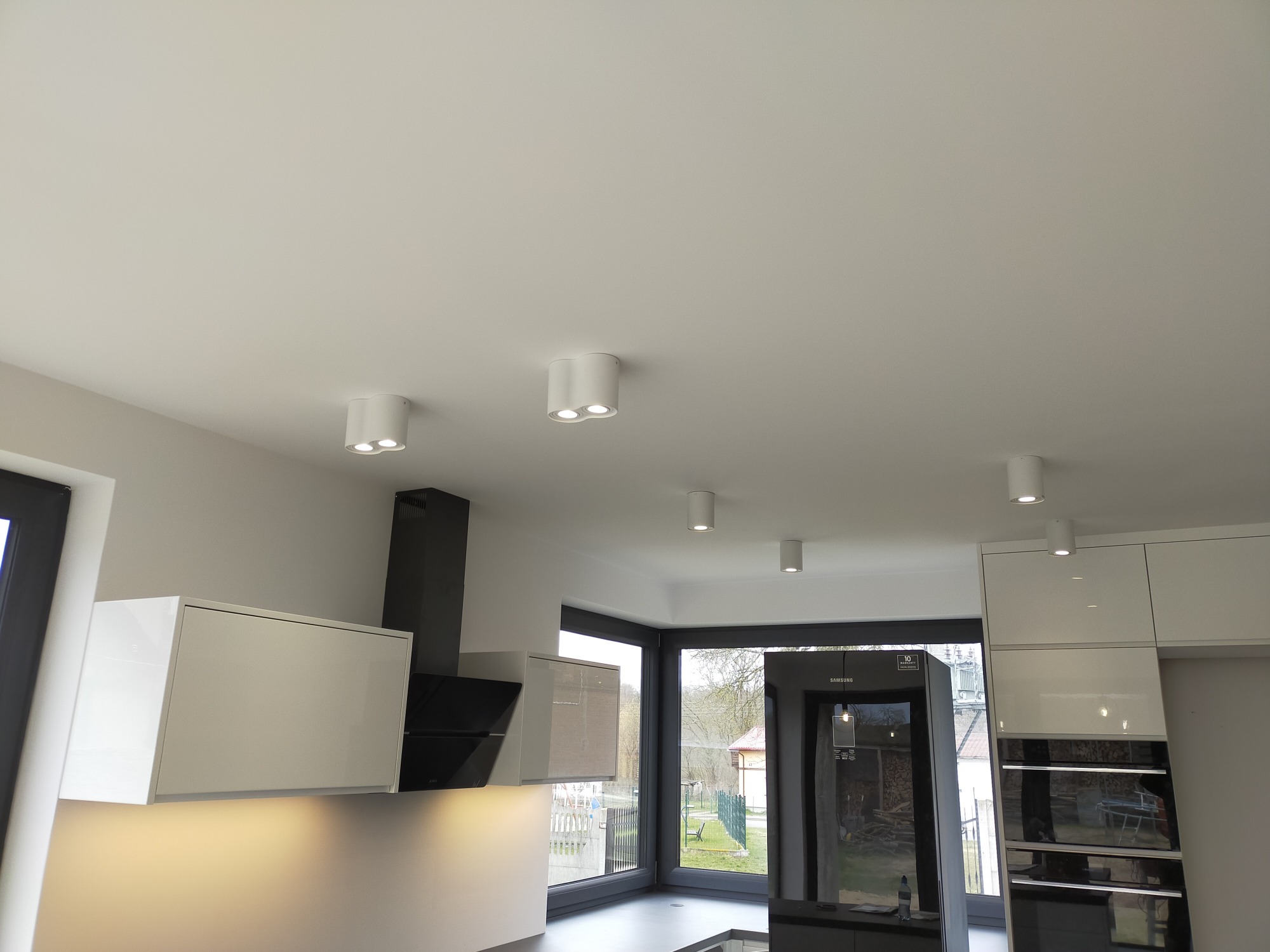 Nowoczesna kuchnia z białymi szafkami i czarnym okapem, oświetlona punktowymi lampami sufitowymi, widok na zewnątrz przez duże okno.