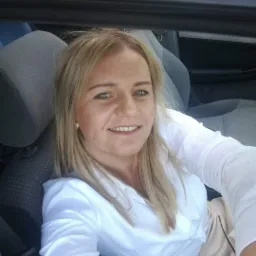 Portret uśmiechniętej blondynki w białej koszuli, siedzącej w samochodzie, prawdopodobnie w drodze na spotkanie z klientem.