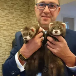 Mężczyzna w garniturze i okularach trzyma dwa brązowe fretki na tle ściany z dekoracyjnej cegły.