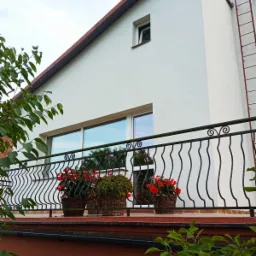 Biały dom z balkonem ozdobionym czarną, falistą balustradą i trzema koszami z czerwonymi kwiatami, widoczne po zewnętrznej stronie, po umyciu.