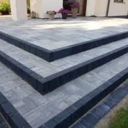 Schody zewnętrzne wyłożone szarą kostką brukową z ciemnymi obrzeżami, prowadzące do jasnego budynku z białymi kolumnami. Widoczne rośliny w donicach.