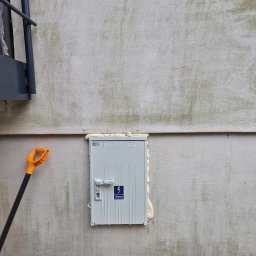 Patryk Szwiergiel - Szara skrzynka elektryczna zamontowana na szarej, surowej ścianie, uszczelniona pianką montażową. Obok, po lewej stronie, widoczny fragment drabiny i łopata z pomarańczową rączką. U dołu gruz...