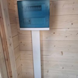 Patryk Szwiergiel - Skrzynka rozdzielcza z niebieską szybką i białym panelem montażowym, zamontowana na ścianie z jasnych desek, widoczny fragment podłogi z materiałem budowlanym.