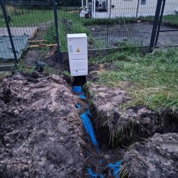 Patryk Szwiergiel - Biała skrzynka elektryczna z symbolem ostrzegawczym, wystająca z ziemi nad wykopem z niebieską rurą osłonową kabla, widok z góry, na tle ogrodzenia z siatki i domku letniskowego.