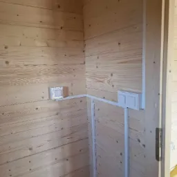 Wnętrze drewnianego domku z widocznymi, nowo zamontowanymi, białymi puszkami i listwami instalacyjnymi podtynkowymi, umieszczonymi na ścianach z boazerii.