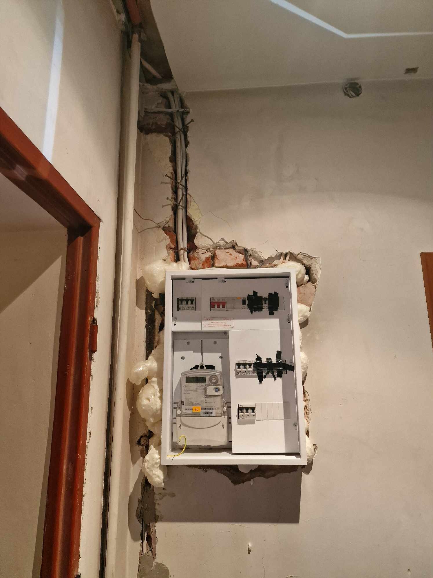 Montaż rozdzielnicy elektrycznej w trakcie prac wykończeniowych; widoczna biała obudowa z zabezpieczeniami, licznikiem i przewodami, osadzona w ścianie z ubytkami, wypełnionymi pianką montażową.