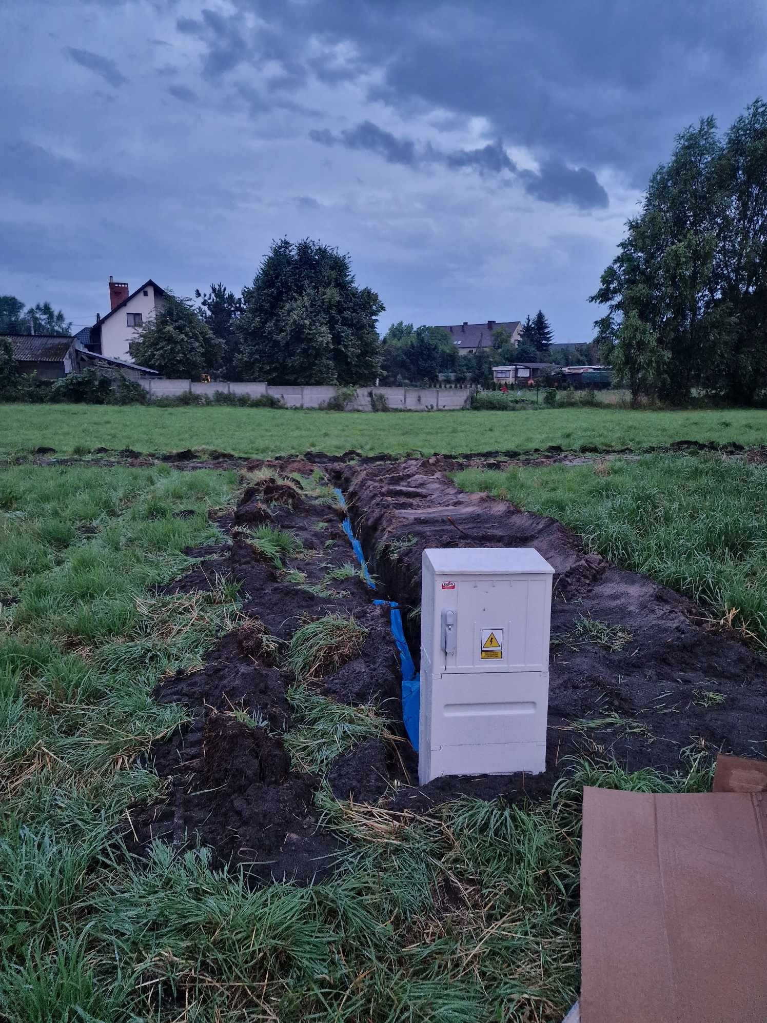 Biała skrzynka elektryczna z symbolem ostrzegawczym na tle wykopu z niebieskimi rurami kablowymi, widok na posesję z domem i pochmurne niebo.