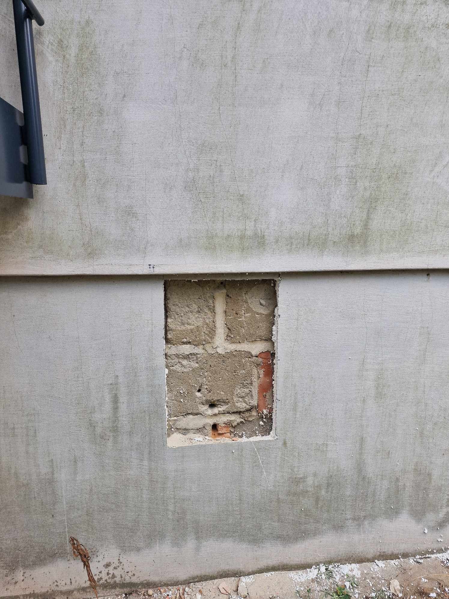 Fragment szarej ściany z otworem odsłaniającym cegły i zaprawę murarską, widoczne ślady wilgoci na powierzchni ściany.