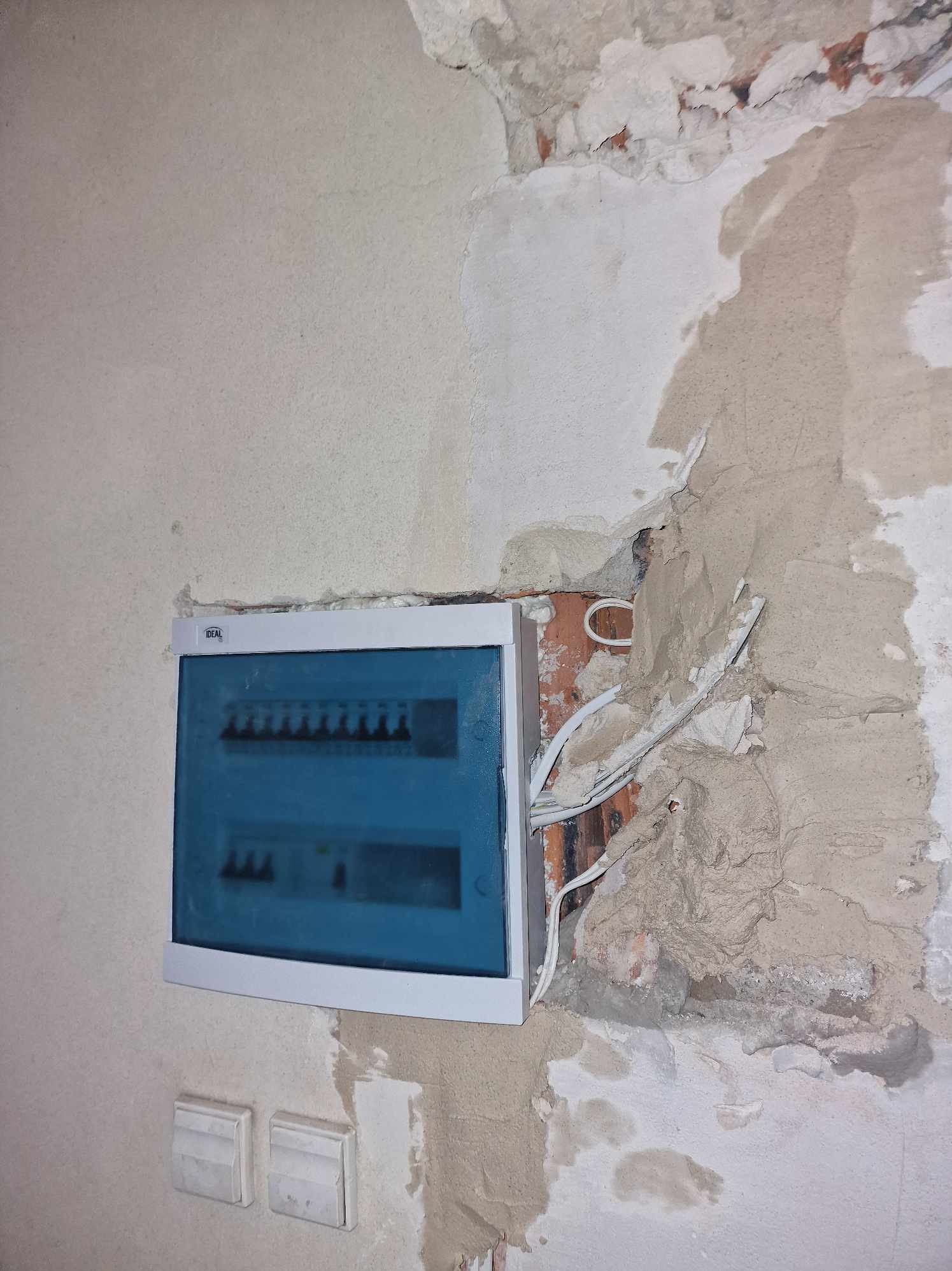 Niebieska skrzynka rozdzielcza z białymi kablami wystającymi ze ściany, częściowo otynkowanej, z widoczną cegłą.