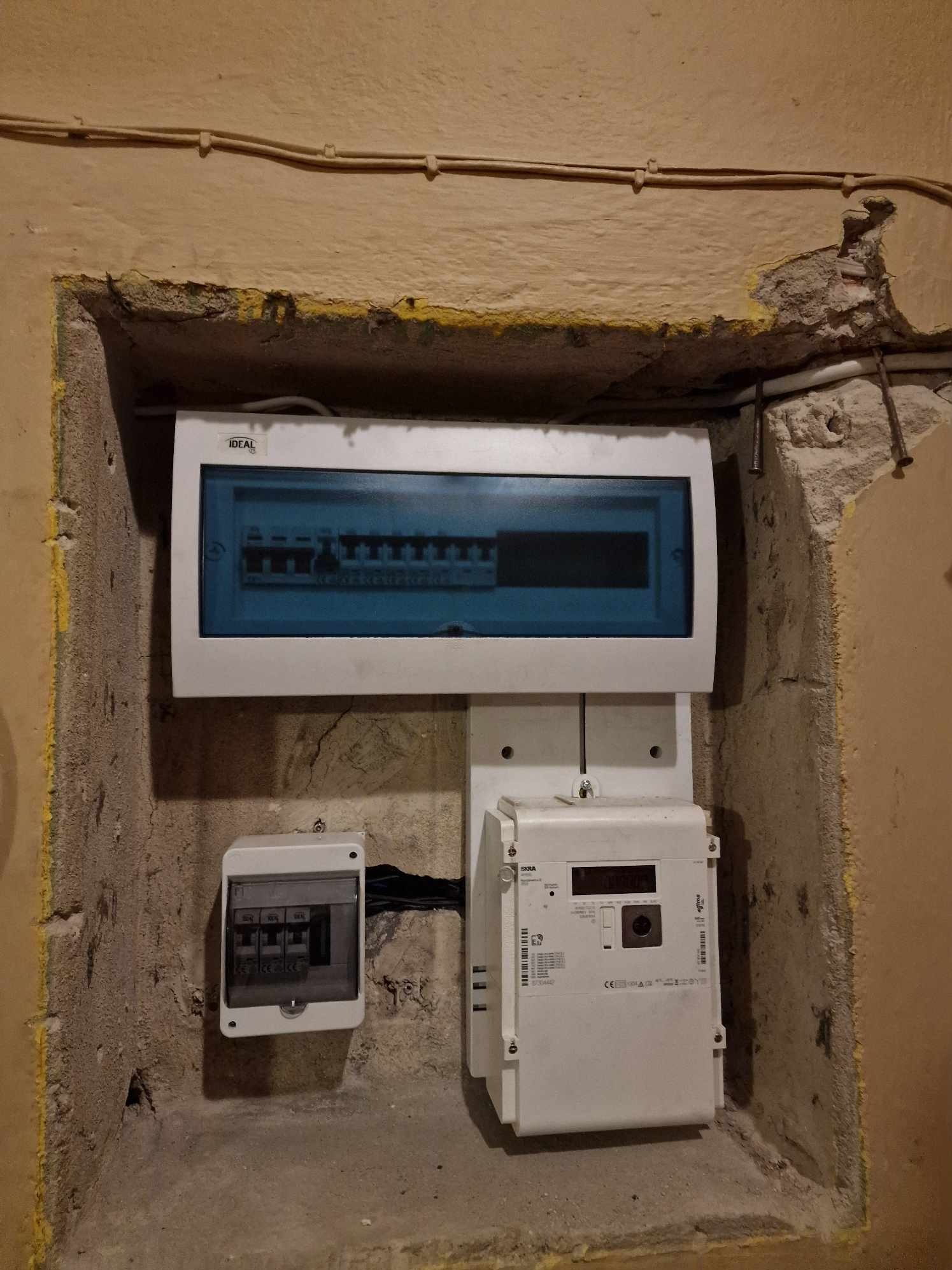 Zamontowana skrzynka rozdzielcza z licznikiem elektrycznym w surowym, otynkowanym wnęce w ścianie, widoczne przewody elektryczne.