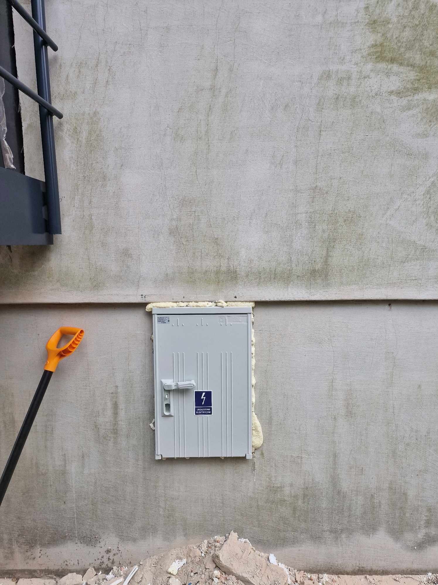 Szara skrzynka elektryczna zamontowana na szarej, surowej ścianie, uszczelniona pianką montażową. Obok, po lewej stronie, widoczny fragment drabiny i łopata z pomarańczową rączką. U dołu gruz...