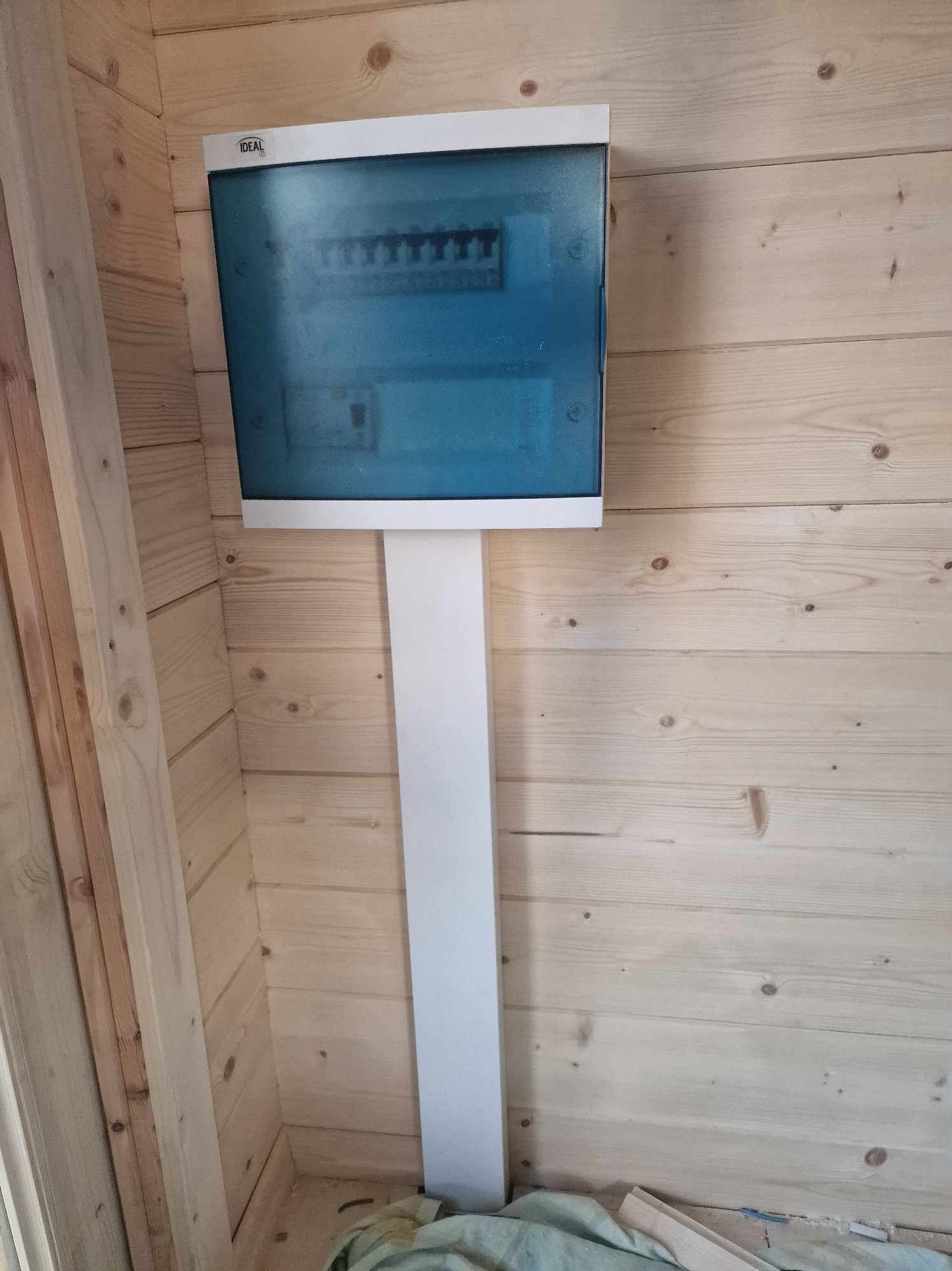 Skrzynka rozdzielcza z niebieską szybką i białym panelem montażowym, zamontowana na ścianie z jasnych desek, widoczny fragment podłogi z materiałem budowlanym.