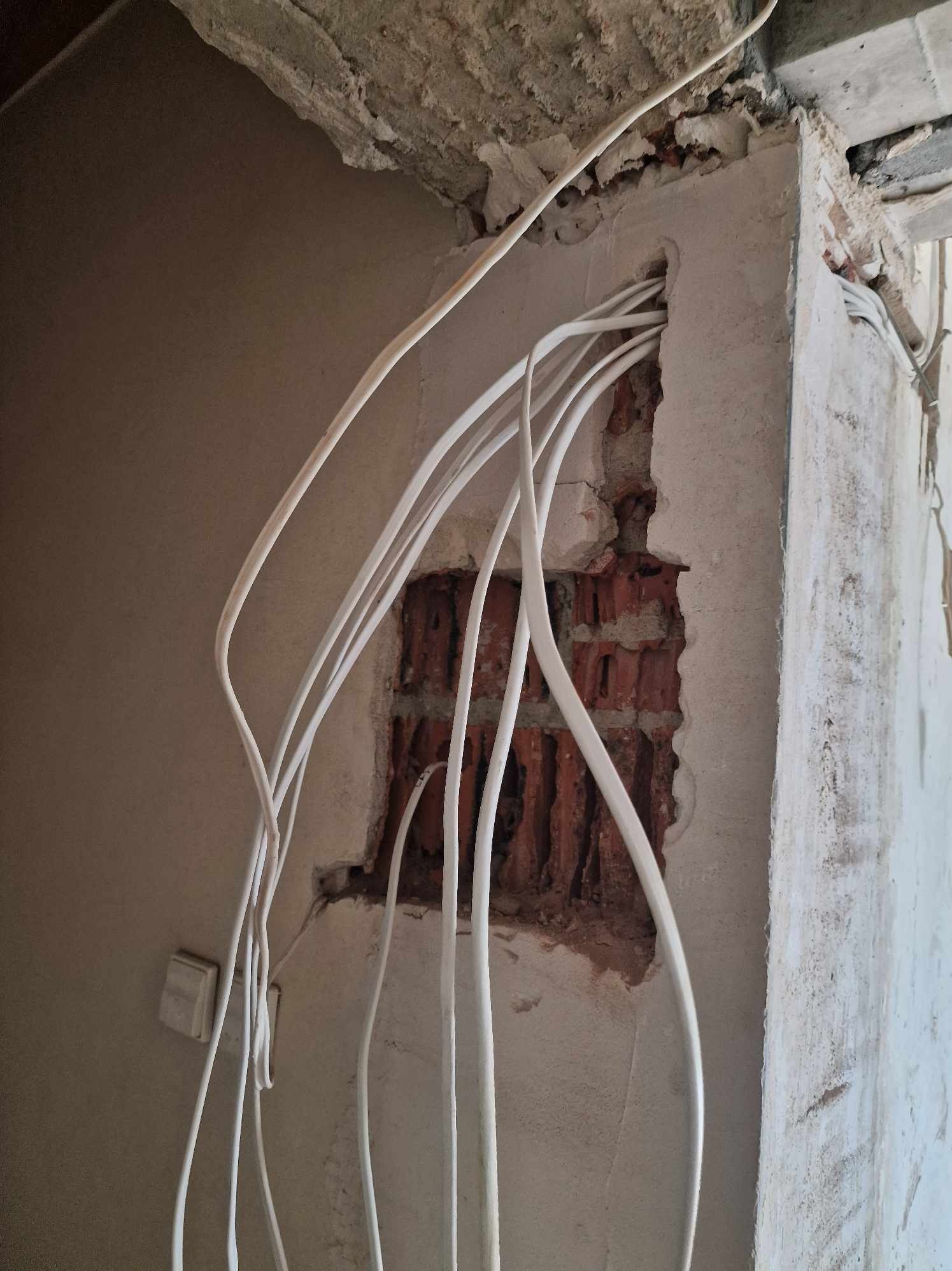 Ściana z odsłoniętą cegłą i wyprowadzonymi przewodami elektrycznymi w białej izolacji, widoczny włącznik światła na ścianie, surowy stan wykończenia.