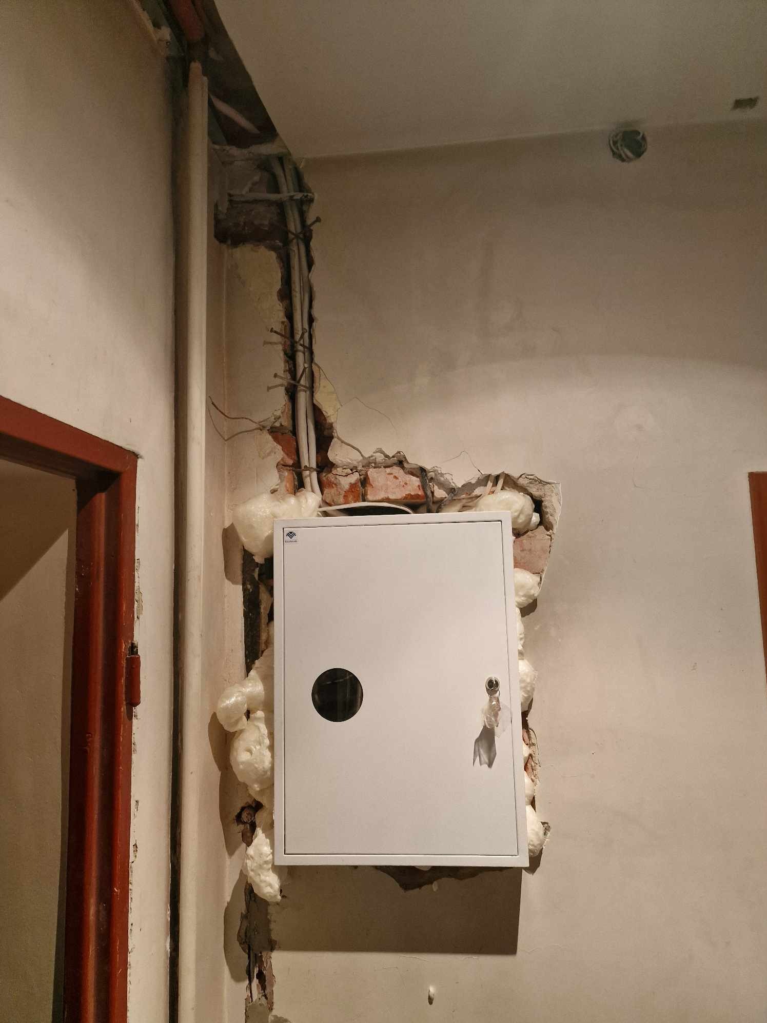 Biała skrzynka elektryczna zamontowana w ścianie z widocznymi uszkodzeniami i pianką montażową, obok pionu z kablami.