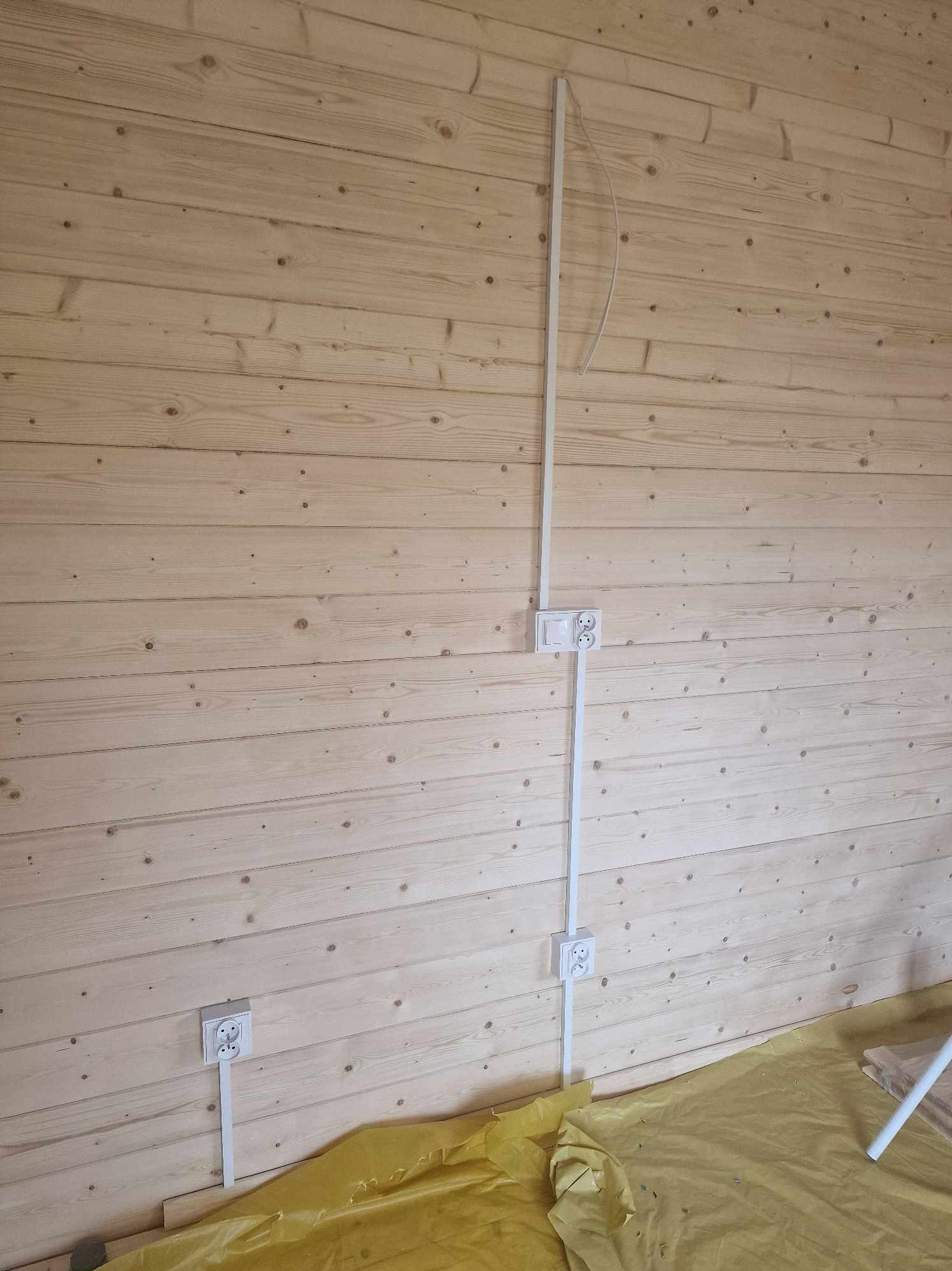 Świeżo położona boazeria, na której zamontowano białe gniazdka i włącznik, a także białe listwy maskujące przewody elektryczne. Podłoga zabezpieczona żółtą folią malarską.