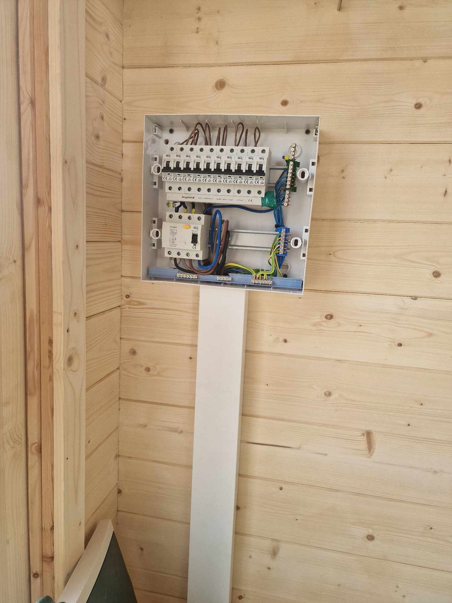 Otwarta skrzynka rozdzielcza z zamontowanymi bezpiecznikami i okablowaniem, umieszczona na drewnianej ścianie wykończonej boazerią.