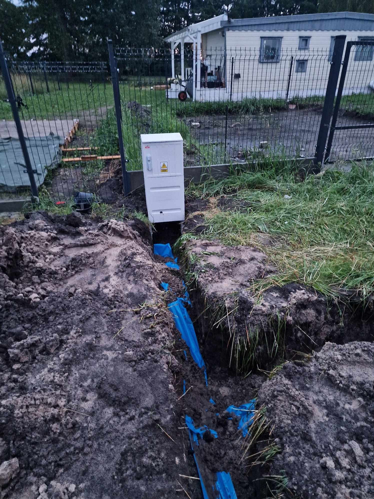 Biała skrzynka elektryczna z symbolem ostrzegawczym, wystająca z ziemi nad wykopem z niebieską rurą osłonową kabla, widok z góry, na tle ogrodzenia z siatki i domku letniskowego.