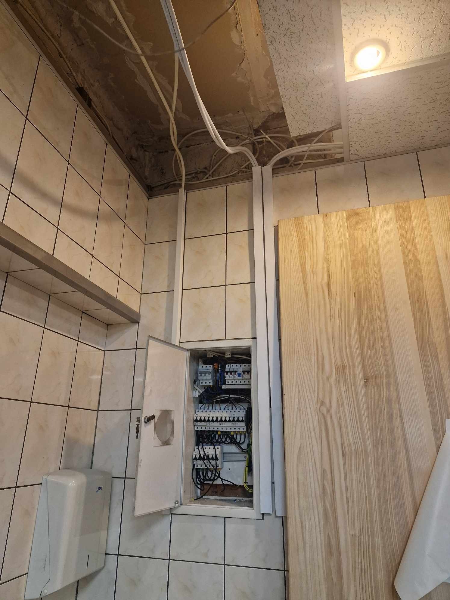 Otwarta skrzynka elektryczna z widocznymi bezpiecznikami i okablowaniem, umieszczona na kafelkowanej ścianie w pomieszczeniu z widocznymi przewodami pod sufitem i suszarką do rąk po lewej stronie.