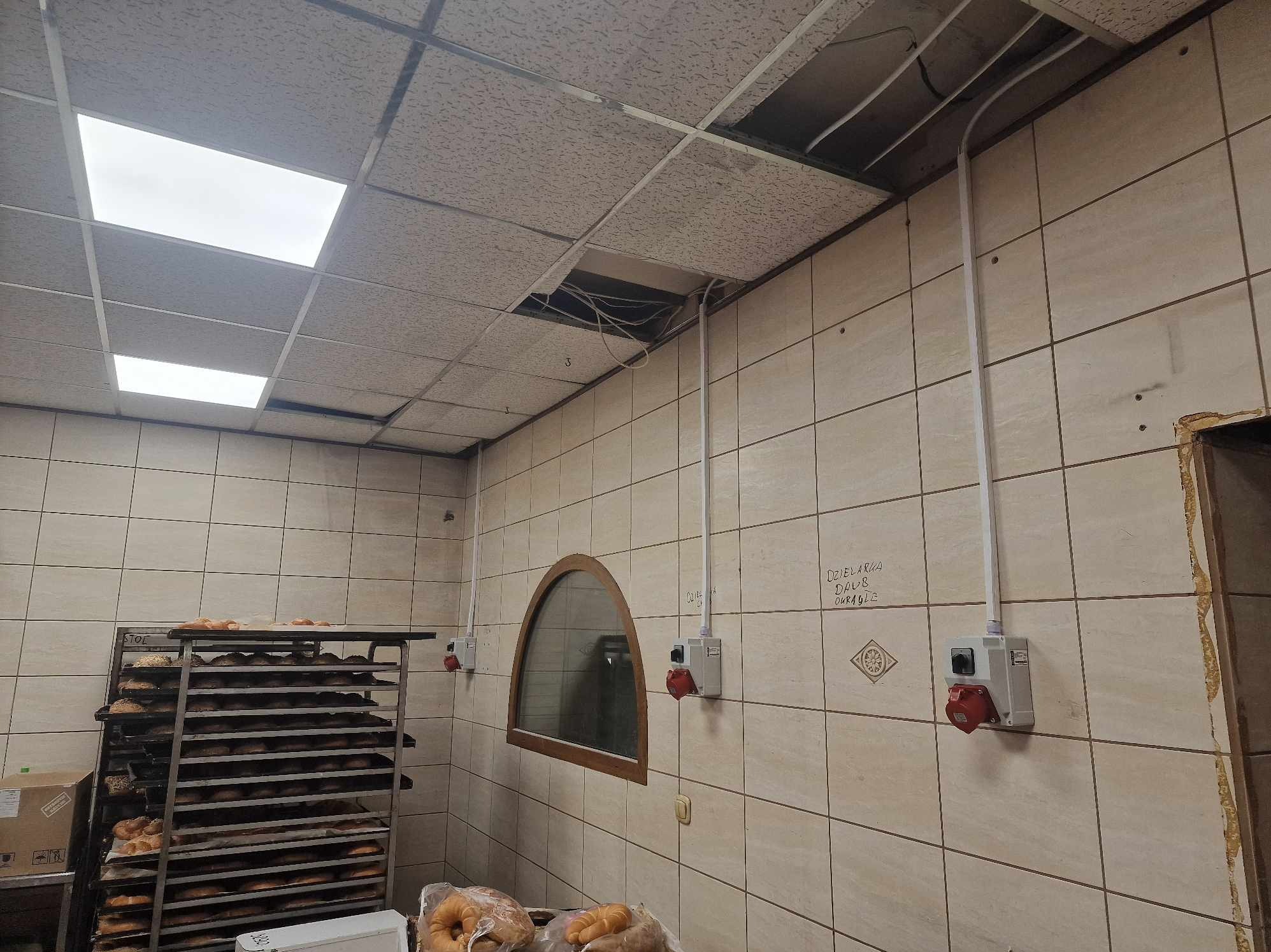 Wnętrze piekarni w trakcie remontu: widoczne odkryte przewody w suficie podwieszanym, wyprowadzone kable w białych rurkach na ścianie z ceramicznych płytek, wózek z pieczywem w tle.