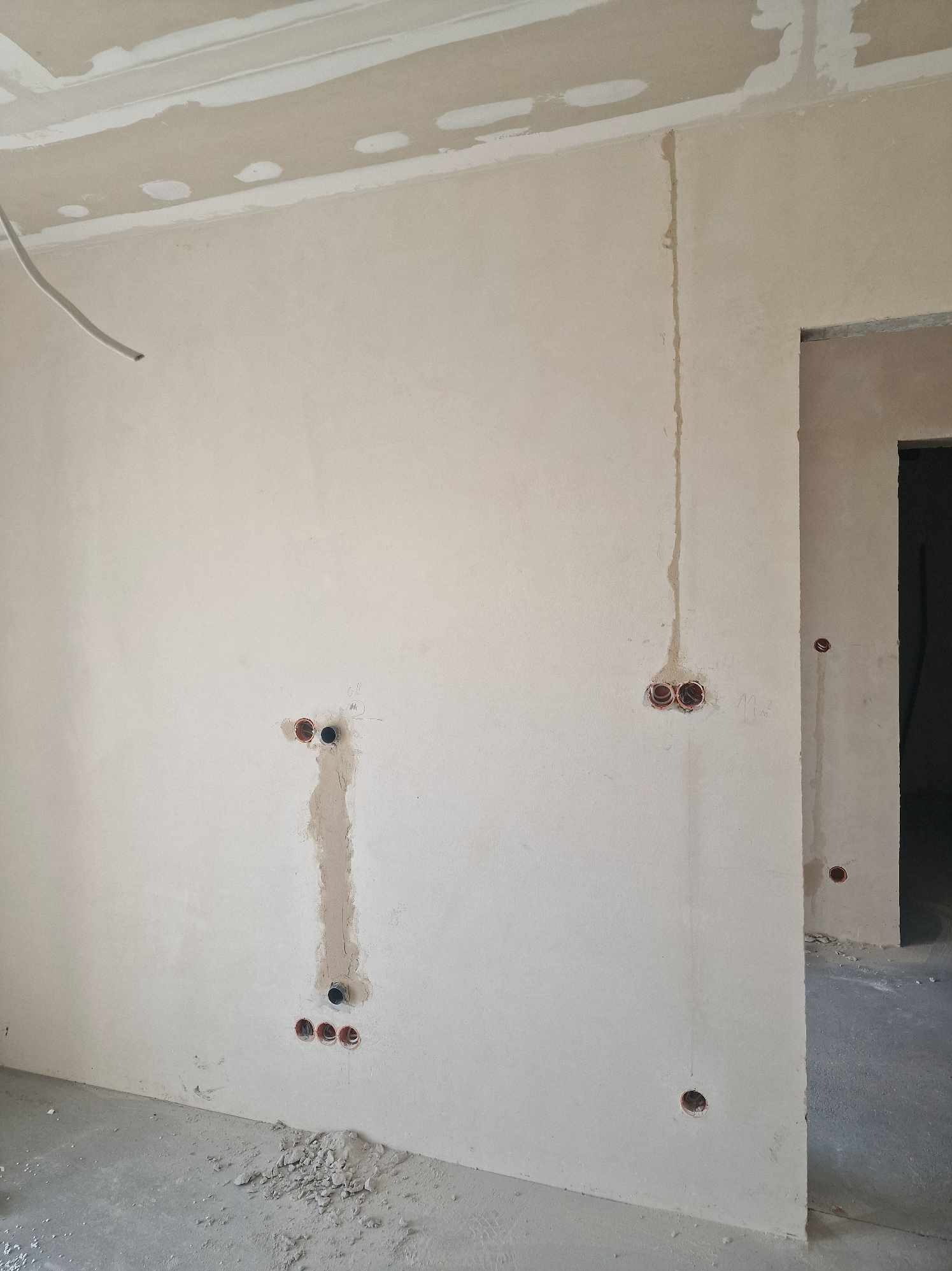 Surowa ściana z wyfrezowanymi otworami pod gniazdka elektryczne i bruzdami pod przewody, widoczne fragmenty przewodów, na podłodze gruz i resztki gipsu.