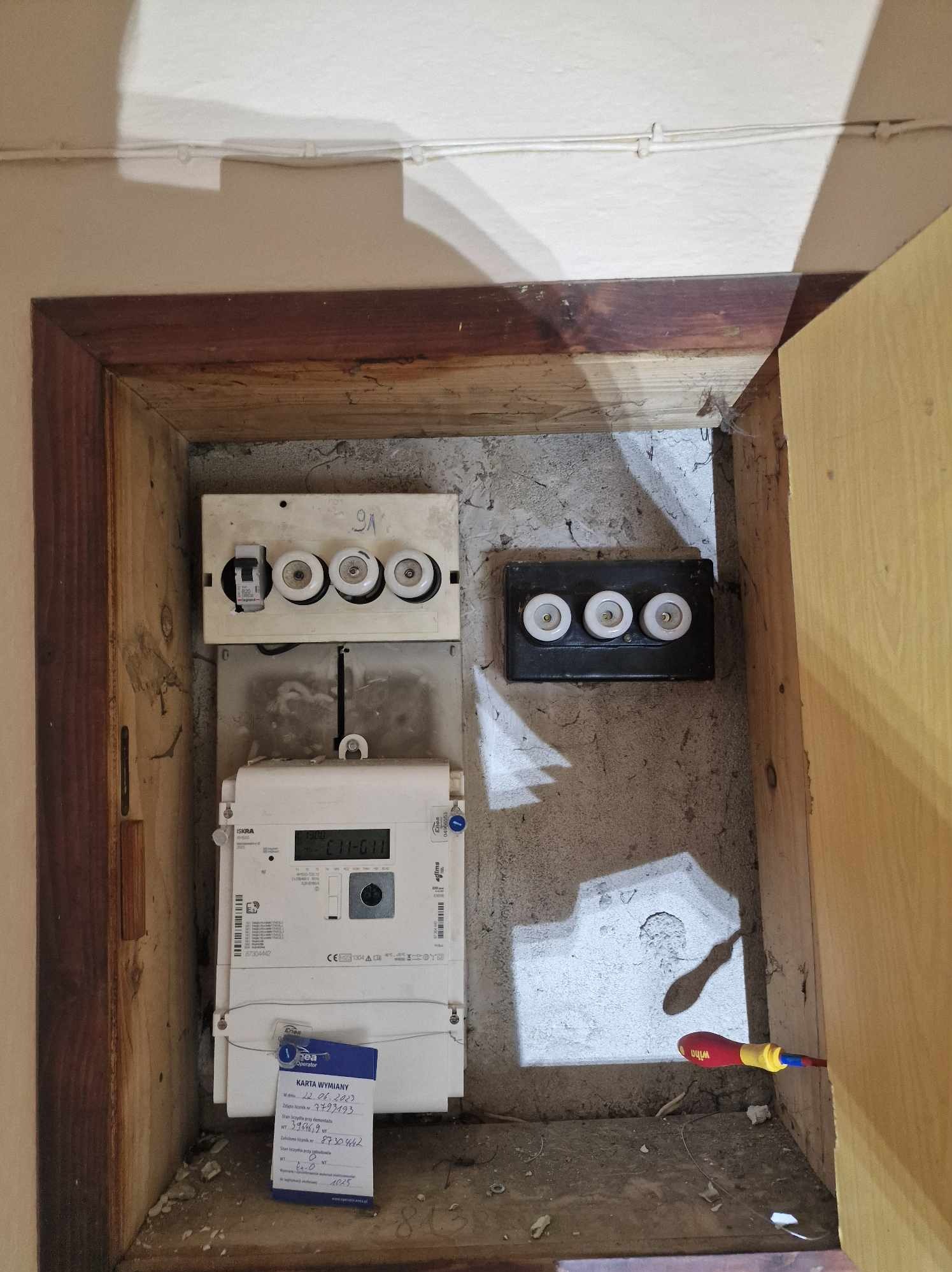 Wnęka z zamontowanym licznikiem elektrycznym, bezpiecznikami topikowymi i dokumentem wymiany z datą 22.06.2007, obok leży śrubokręt Wiha.