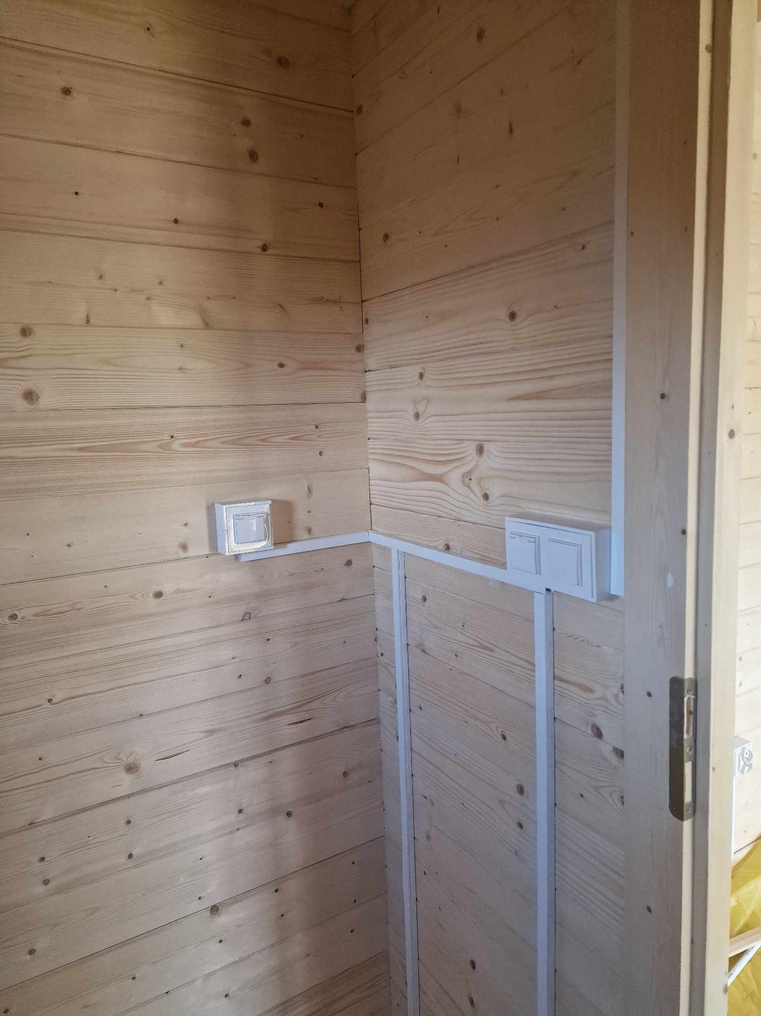 Wnętrze drewnianego domku z widocznymi, nowo zamontowanymi, białymi puszkami i listwami instalacyjnymi podtynkowymi, umieszczonymi na ścianach z boazerii.