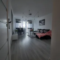 Jasne wnętrze mieszkania z szarą podłogą, widok zza otwartych drzwi, w tle stół z krzesłami, sofa z poduszkami i różowym pledem oraz wiszący fotel typu huśtawka.