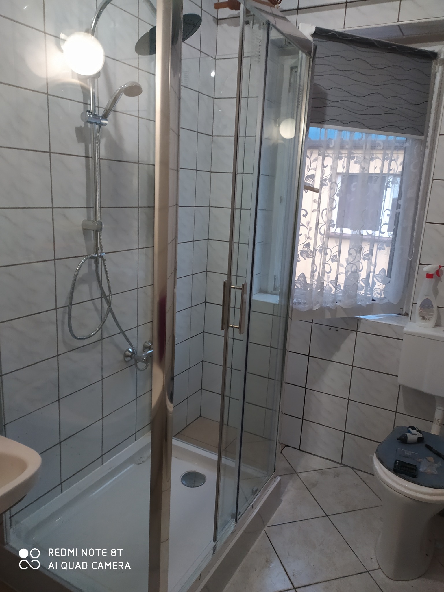 Kabina prysznicowa z chromowanymi profilami i białym brodzikiem w łazience wyłożonej kwadratowymi, białymi płytkami. Widoczny zestaw prysznicowy z deszczownicą. Na sedesie leżą narzędzia.