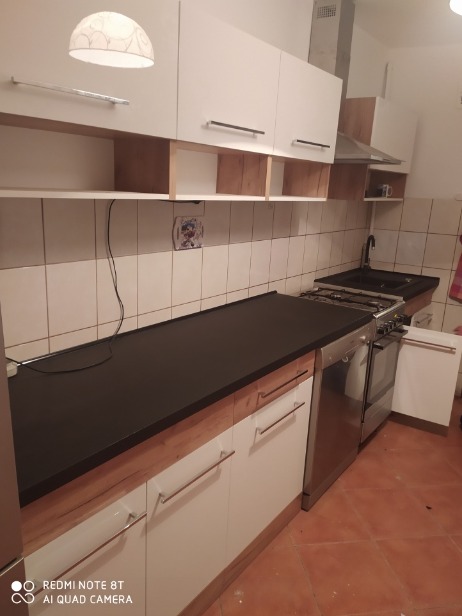 Nowoczesna kuchnia z białymi szafkami, drewnianymi akcentami i czarnym blatem, widok na kuchenkę gazową i okap, płytki na ścianie.