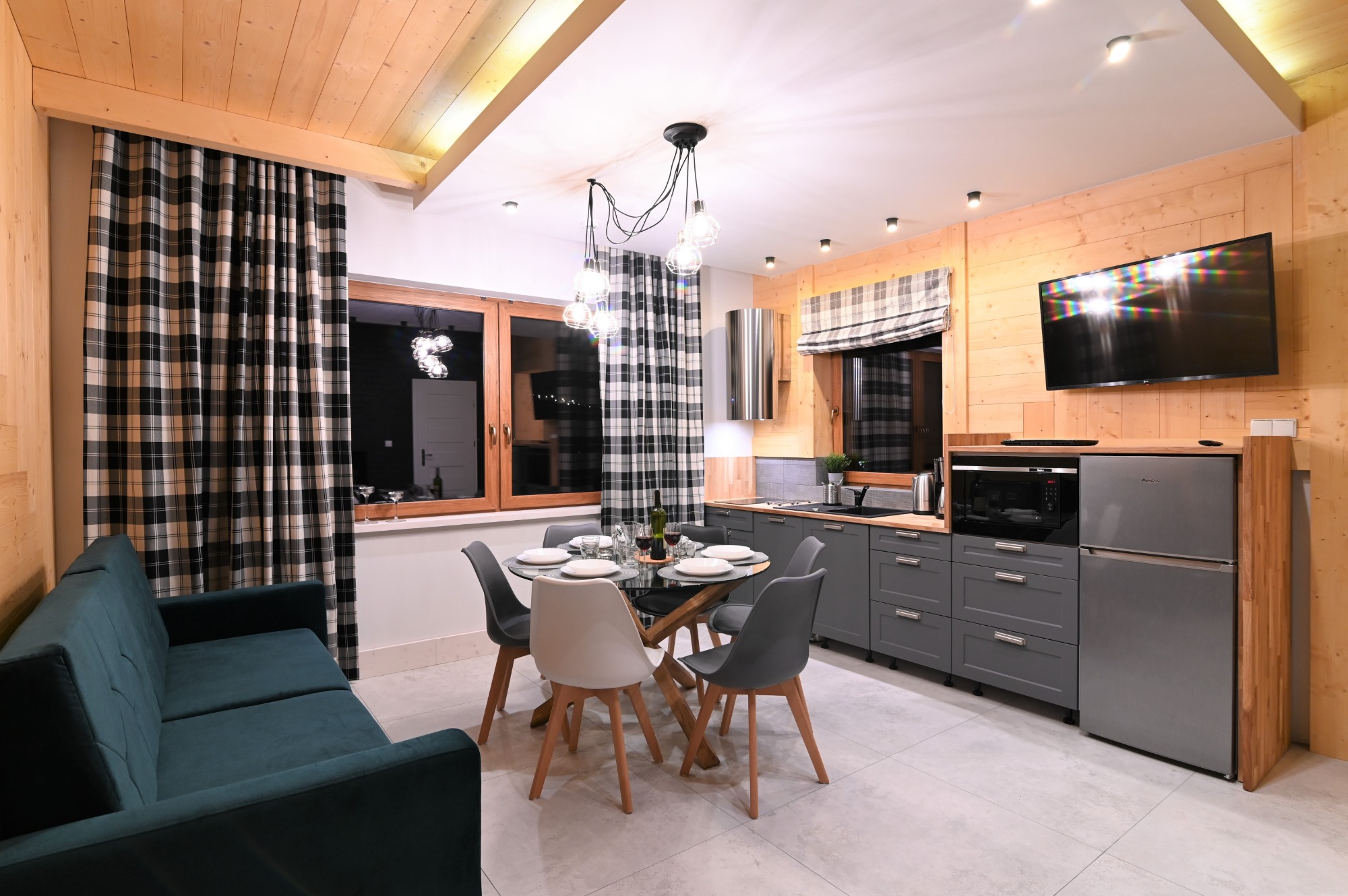 Jasne wnętrze apartamentu w stylu góralskim: drewniane ściany, szare meble kuchenne, okrągły stół z nakryciem, zielona sofa i okna z czarno-białymi zasłonami w kratę.
