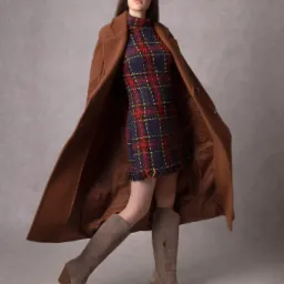 Modelka w ruchu, prezentująca tweedową sukienkę w kratę, brązowy płaszcz i zamszowe kozaki na obcasie na neutralnym tle.