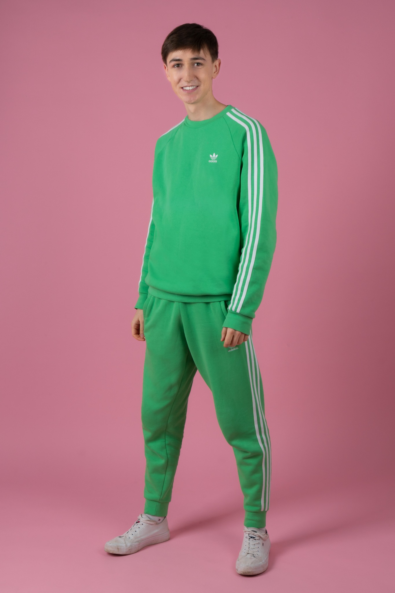 Młody mężczyzna uśmiecha się, ubrany w zielony dres Adidas z trzema białymi paskami, pozuje na różowym tle.