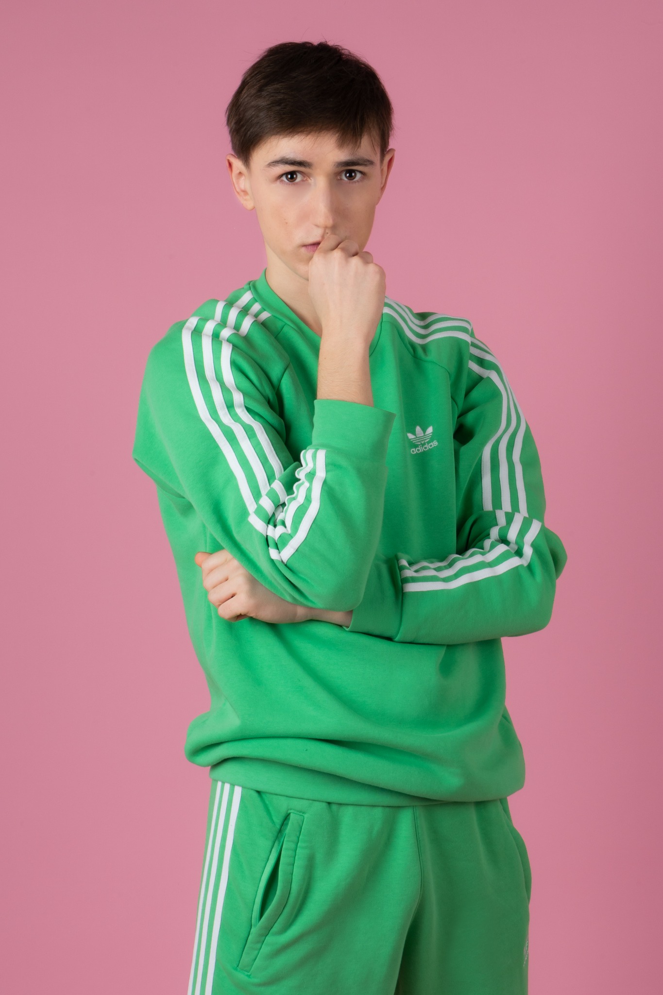 Młody mężczyzna w zielonym dresie Adidas z trzema białymi paskami, pozujący na różowym tle, zamyślony wyraz twarzy.