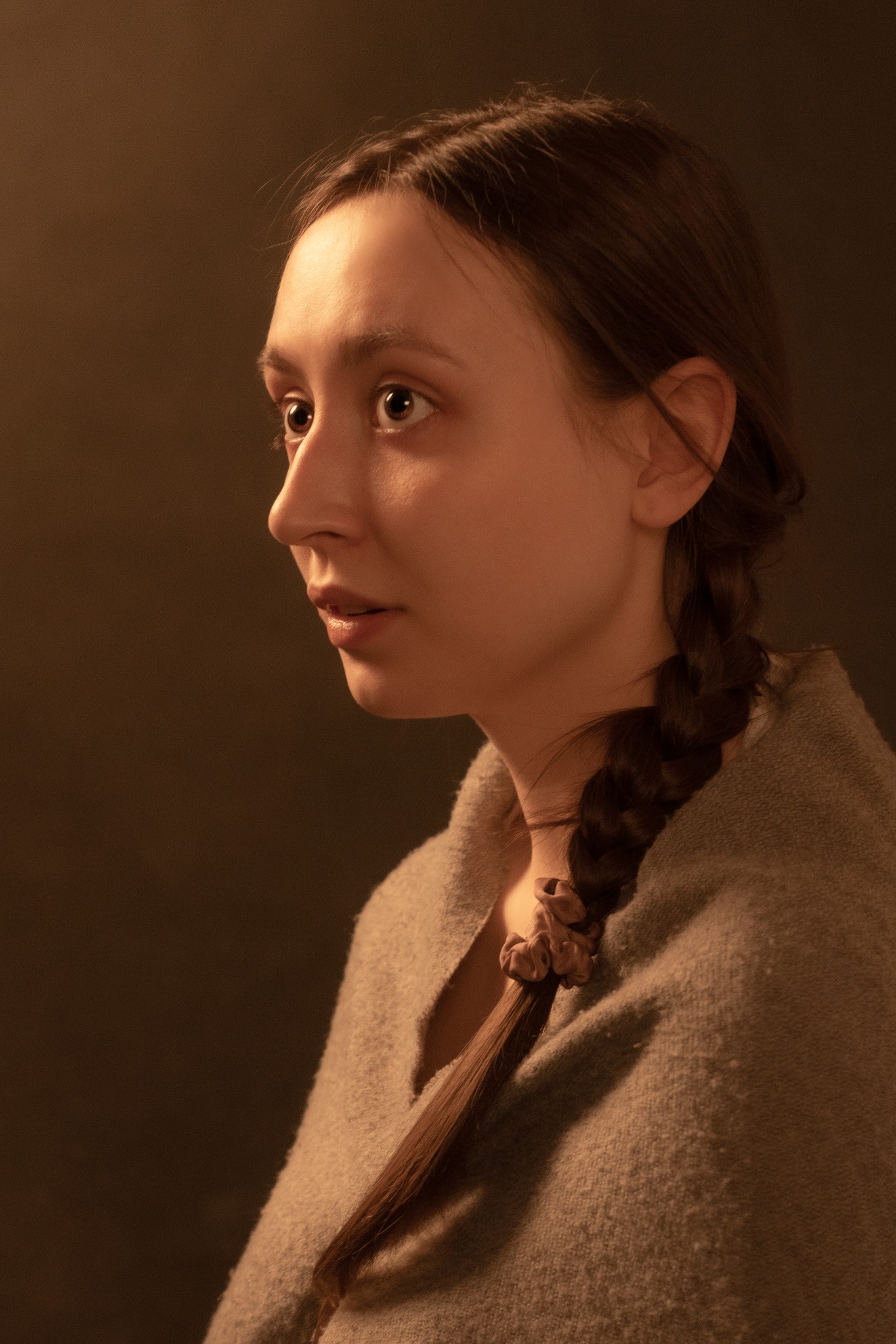 Portret młodej kobiety z warkoczem, profil, miękkie światło, neutralne tło, ubrana w szary sweter.