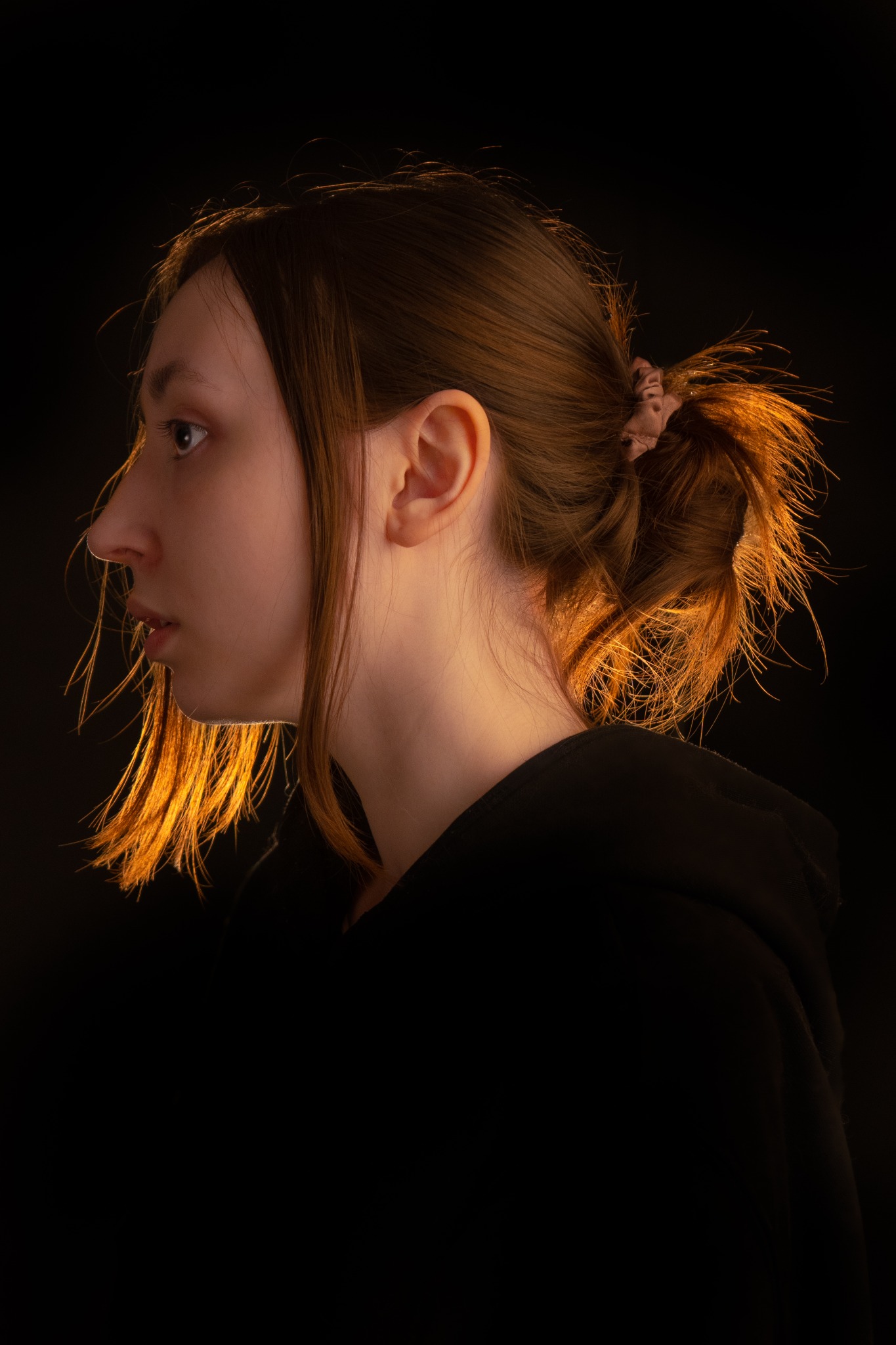 Profil młodej kobiety z włosami upiętymi w kok, oświetlonymi ciepłym światłem na czarnym tle.
