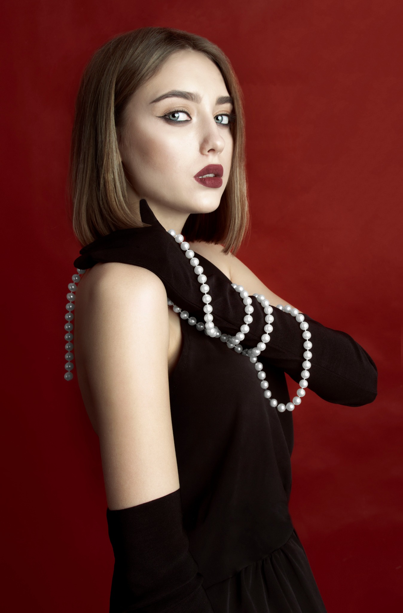 Portret kobiety w czarnej sukience i długich rękawiczkach, z perłami na ramieniu, na czerwonym tle, z wyrazistym makijażem oczu i ust.