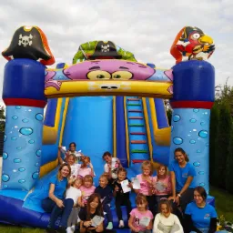 Grupa dzieci i dwie kobiety w niebieskich koszulkach siedzą przed dmuchanym zamkiem w pirackim stylu, trzymając dyplomy na trawie.