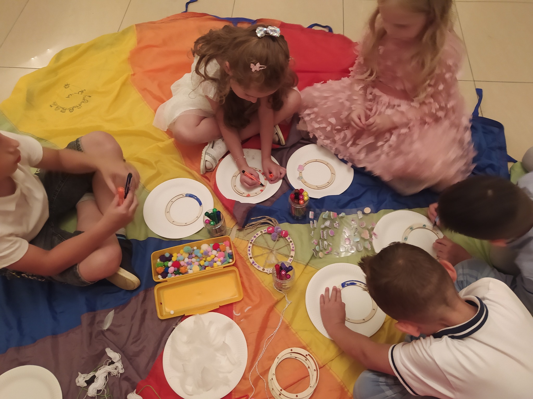 Dzieci siedzą na kolorowej płachcie i dekorują papierowe talerze kolorowymi pisakami, pomponami i naklejkami, tworząc ozdoby.