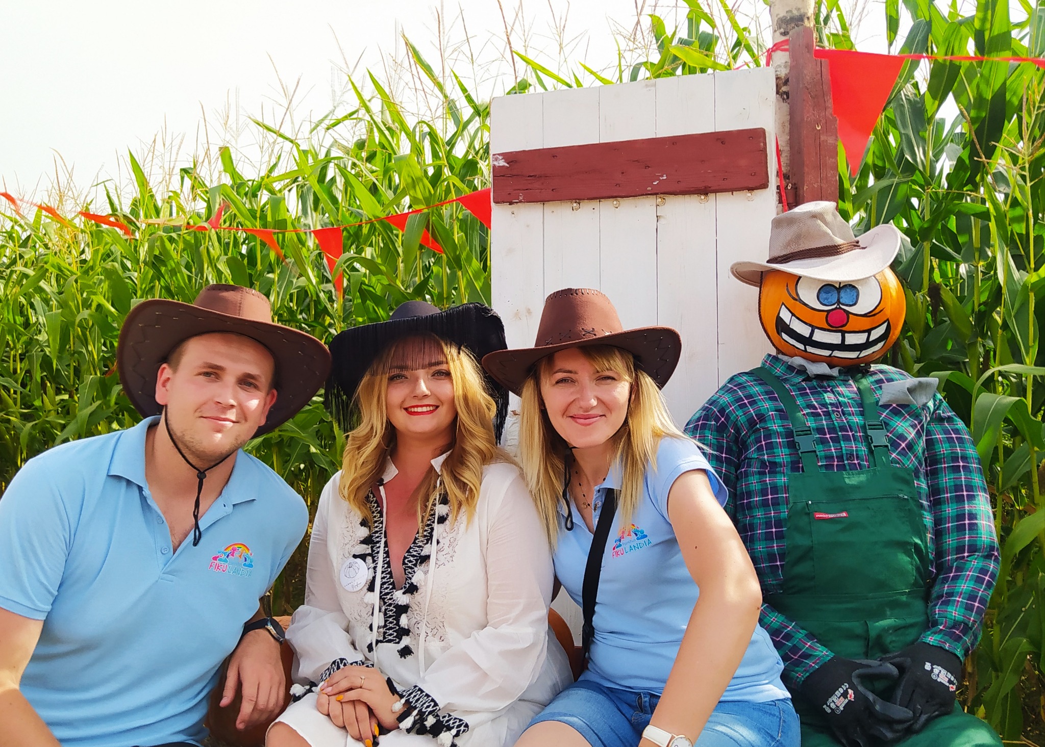 Trzy osoby w kowbojskich kapeluszach siedzą obok dyniowego stracha na tle labiryntu kukurydzy z dekoracjami i logo firmy na koszulkach.