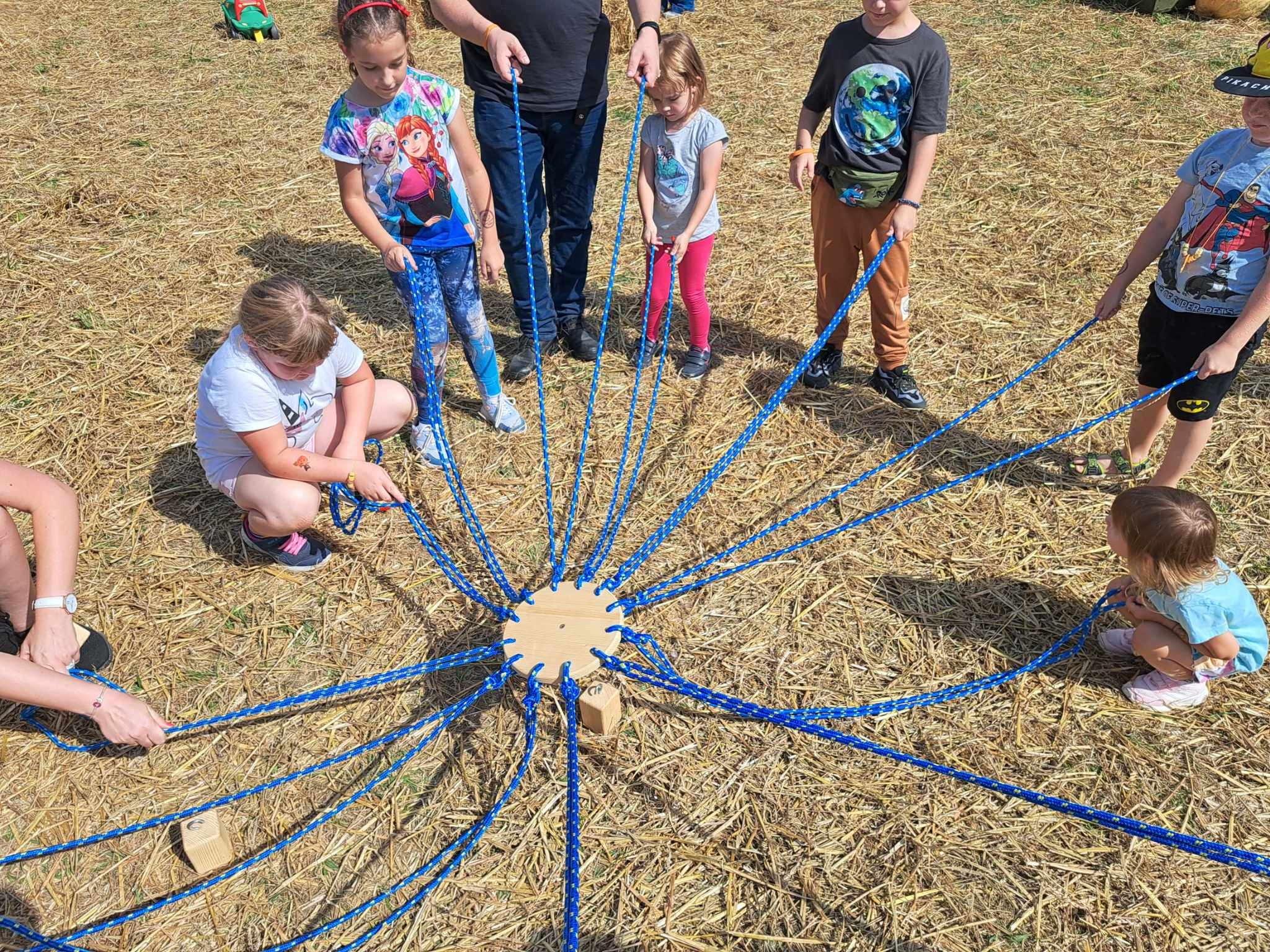 Grupa dzieci i dorosłych uczestniczy w grze zespołowej na trawiastym terenie, trzymając niebieskie liny przymocowane do drewnianego dysku z blokami.