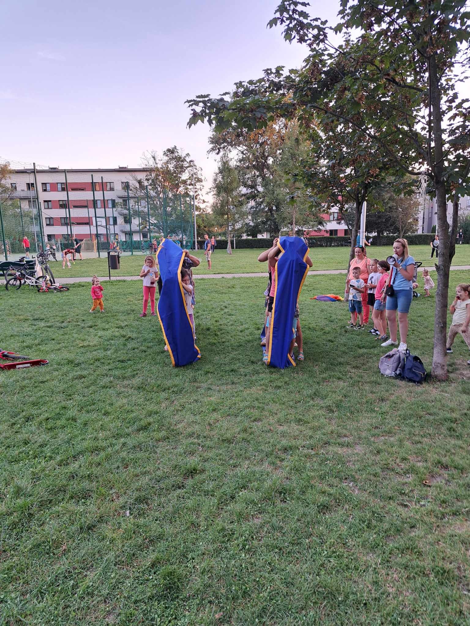 Przygotowania do animacji dla dzieci w Zielonkach: rozkładanie dużych, niebieskich tub z żółtymi brzegami na trawie, w tle grupa dzieci i dorosłych oraz blok mieszkalny.