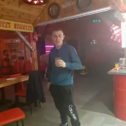 Mężczyzna w dresie Calvin Klein trzyma szklankę z napojem w barze urządzonym w stylu warsztatu samochodowego, z oponą jako żyrandolem i dekoracjami motoryzacyjnymi.