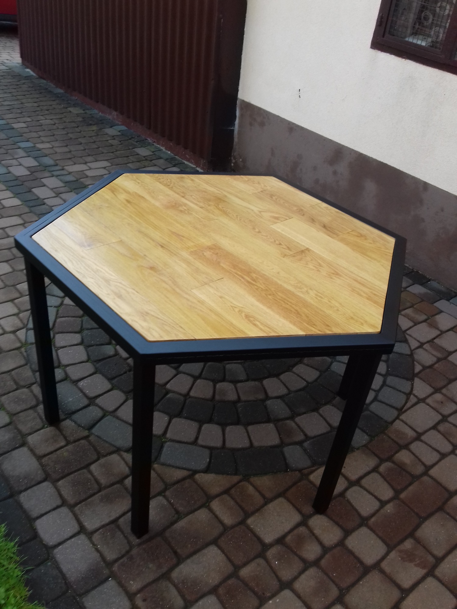 Stół o heksagonalnym blacie z jasnego drewna i czarną, metalową ramą, stojący na kostce brukowej.