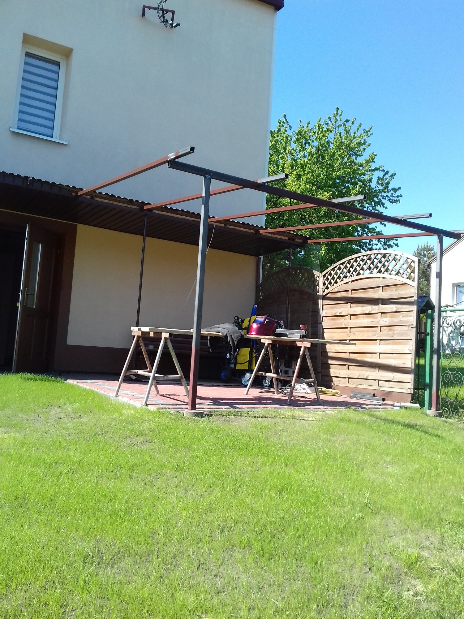 Stalowa konstrukcja z zadaszeniem w trakcie montażu przy budynku mieszkalnym, widoczne narzędzia i elementy ogrodzenia