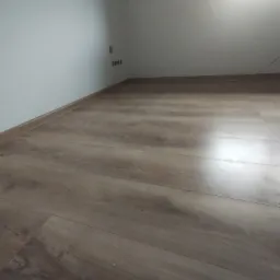 Wykończone wnętrze poddasza z panelami podłogowymi w odcieniach brązu, widoczna biała ściana z gniazdkami elektrycznymi i listwa przypodłogowa.